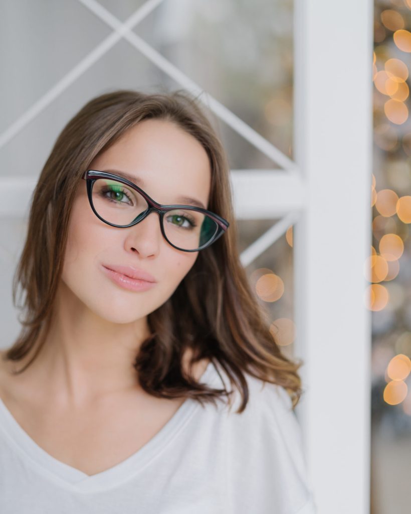 Vertical shot of charming woman in optical glasses, dressed casually, poses indoor against Christams tree, has healthy soft skin, enjoys spare time in domestic atmosphere. People and leisure